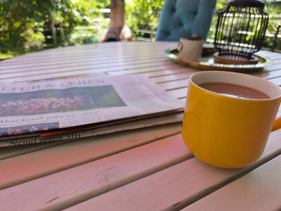 Gelber Becher mit Tee auf einem Gartentisch links liegt eine Tageszeitung,